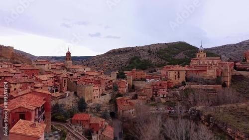 Aerial footage showcasing Albarracin medieval skyline with red stone houses, fortress walls and historic churches set among dramatic cliffs and rugged mountain terrain.