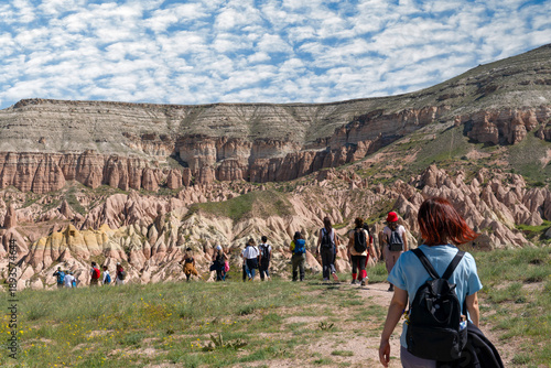 Rear view of a large group of hikers walking in a single line towards massive layered rock formations in Cappadocia, Turkey