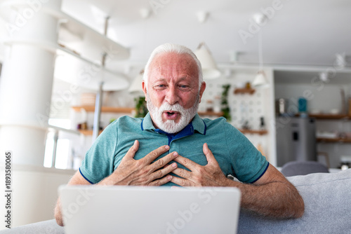 Surprised senior man looking at laptop screen with hands on chest. Shocked elderly male reacting to good news or unexpected results at home. Amazed retired person using computer in living room.