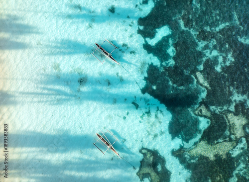 Aerial view of two fishing boats, sea with clear azure water at sunrise in summer. Top drone view of floating boats and palm shadows in transparent ocean. Travel in Zanzibar. Tropical seascape. Exotic