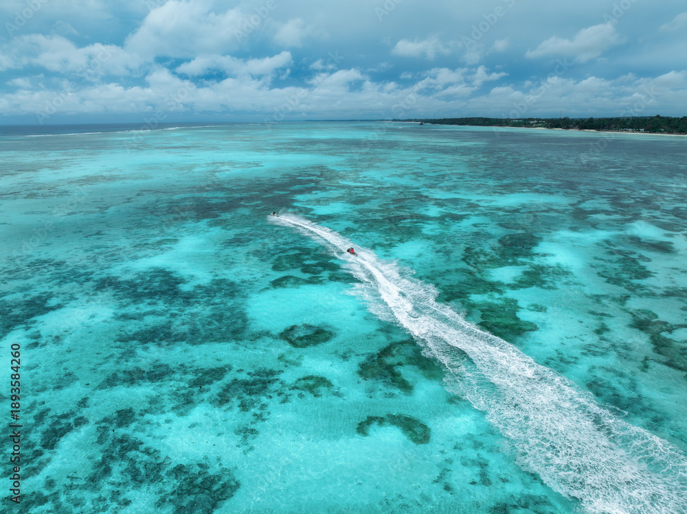Fototapeta premium Aerial view of jet skis over shallow turquoise sea with reef in Zanzibar. Clear tropical water, watercraft trails, summer vacation, coastal travel and marine leisure activity. Top drone view. Exotic