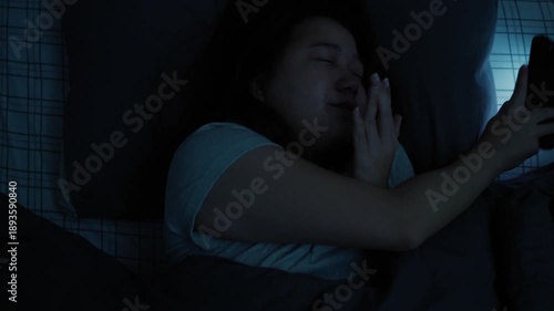 Asian woman unable to sleep, using her smartphone before bedtime and enjoying short-form video content on a platform, with the phone�s light illuminating her face