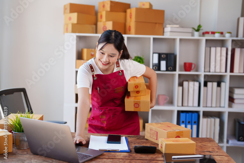 Cuadro en lienzo Young Asian woman entrepreneur in home office holding cardboard boxes and using