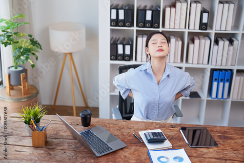 Young woman feeling discomfort and back pain while stretching her lower back at an office desk, illustrating fatigue, stress, and health issues from prolonged sitting