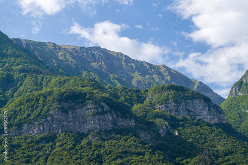 Verdant hillsides with exposed rocky cliffs rise sharply beneath a blue sky scattered with clouds in the Massif des Bauges, French Alps.