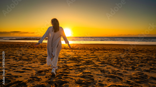 Beautiful middle-aged woman enjoying walk on sandy beach on sunset on windy spring day. Back view