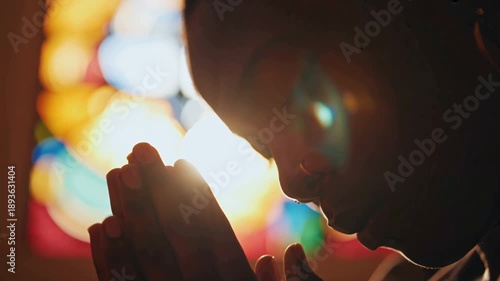 Cinematic 4K stock video, close-up pensive Black woman praying, eyes closed, hands clasped tears glistening, sunlight streaming colorful stained-glass window, delicate lens flare, warm golden hour