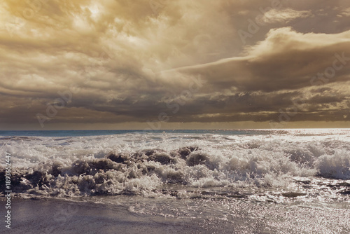 Breaking sea waves and foamy surf on a sandy beach with dramatic storm clouds and blue sky. Scenic seascape background, nature and travel concept.
