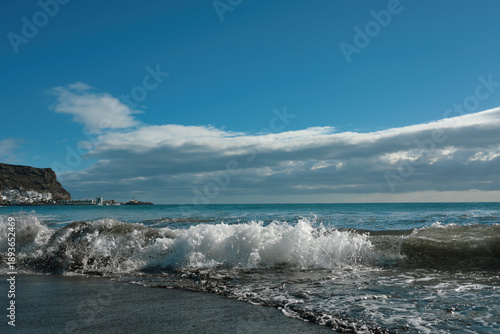 Breaking sea waves and foamy surf on a sandy beach with dramatic storm clouds and blue sky. Scenic seascape background, nature and travel concept.
