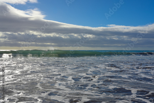 Breaking sea waves and foamy surf on a sandy beach with dramatic storm clouds and blue sky. Scenic seascape background, nature and travel concept.