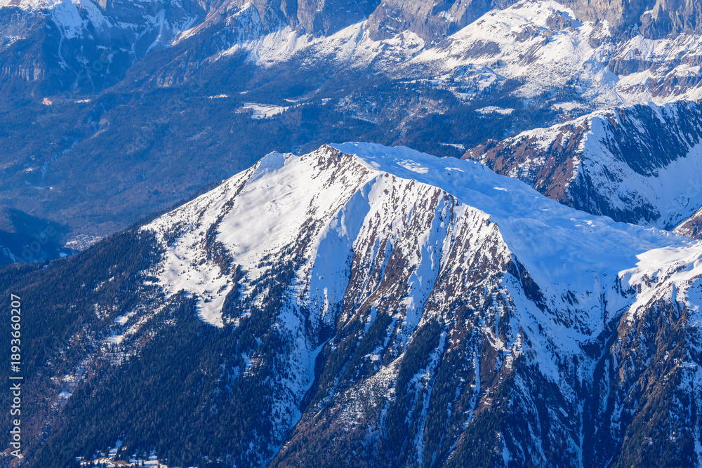 Fototapeta premium High angle view of the Aiguillette des Houches showing a broad snow-covered plateau bordered by steep forested slopes and deep valleys, illuminated by crisp winter sunlight.