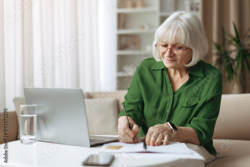 Foto Senior woman writing notes at home while using laptop computer, thoughtful elderly female managing documents, retirement planning, learning and paperwork in a relaxed home environment