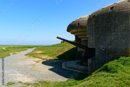 Foto A preserved World War II concrete artillery bunker with a long gun faces the open coastal landscape at Longues-sur-Mer, Normandy, under a clear blue sky and bright sunlight