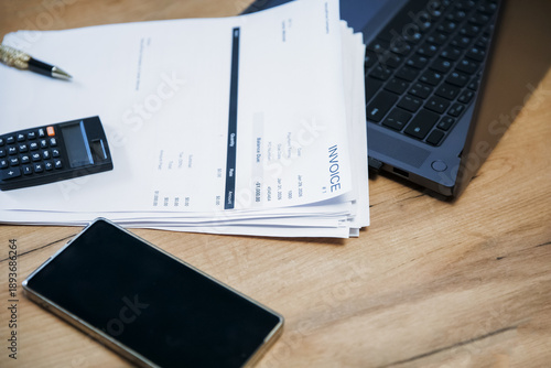 Stack of invoice documents on a wooden desk with a calculator, a pen, a phone, and a laptop. Business accounting concept for financial management.