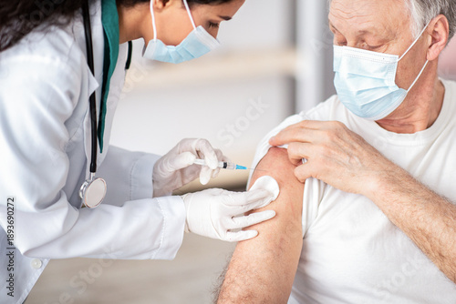 A healthcare worker is giving a vaccine to an older man in a clinic. The man is receiving the shot on his arm while wearing a mask.