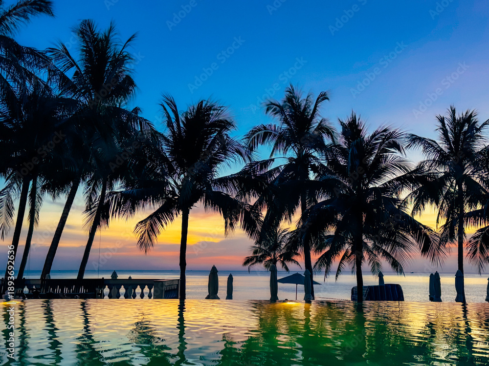 Obraz premium Stunning Sunset at Luxury Beach Resort with Infinity Pool, Silhouetted Palm Trees and Vibrant Twilight Sky in Vietnam