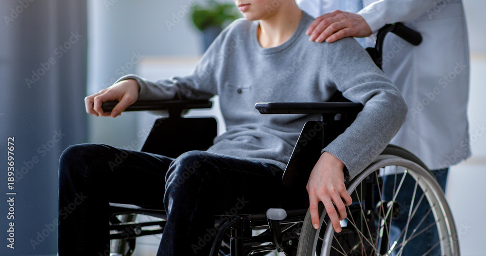 Obraz premium A young boy with light skin is seated in a wheelchair. A healthcare worker is standing beside him, gently placing a hand on his shoulder. They are in a bright clinical environment.