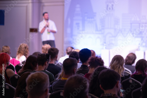 audience sits in conference hall listening to speaker on stage, bright backdrop cityscape displayed, event energy professional mood
