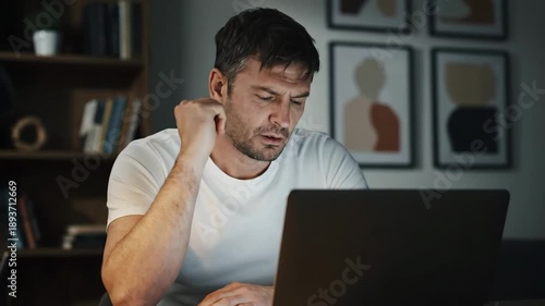 Tired adult caucasian man working late on a laptop at his home office desk. Stressed freelancer concentrating on a difficult project and feeling exhausted from overtime work