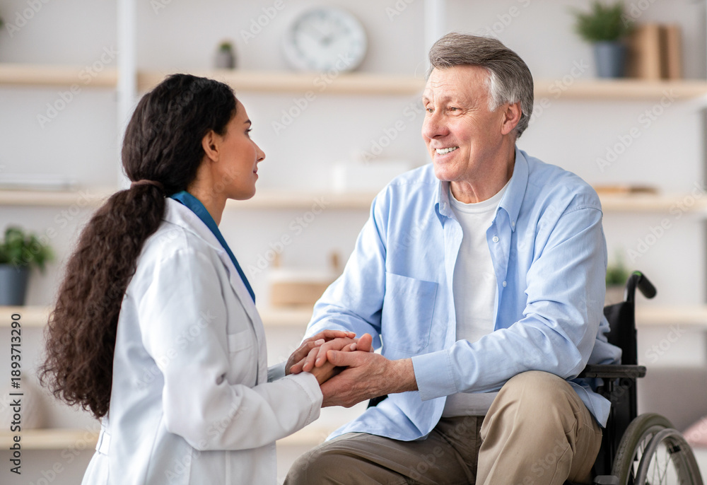 Obraz premium A medical professional talks to a senior man using a wheelchair. They are in a bright therapy room with soft decorations. The atmosphere shows support and care.