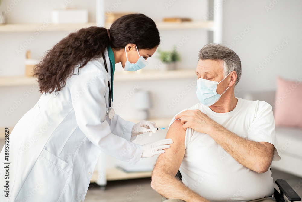 Obraz premium A health worker assists an older man with gray hair in receiving a vaccine. Both are wearing masks, and the setting is a well-lit clinic. The health worker is focused on the task.
