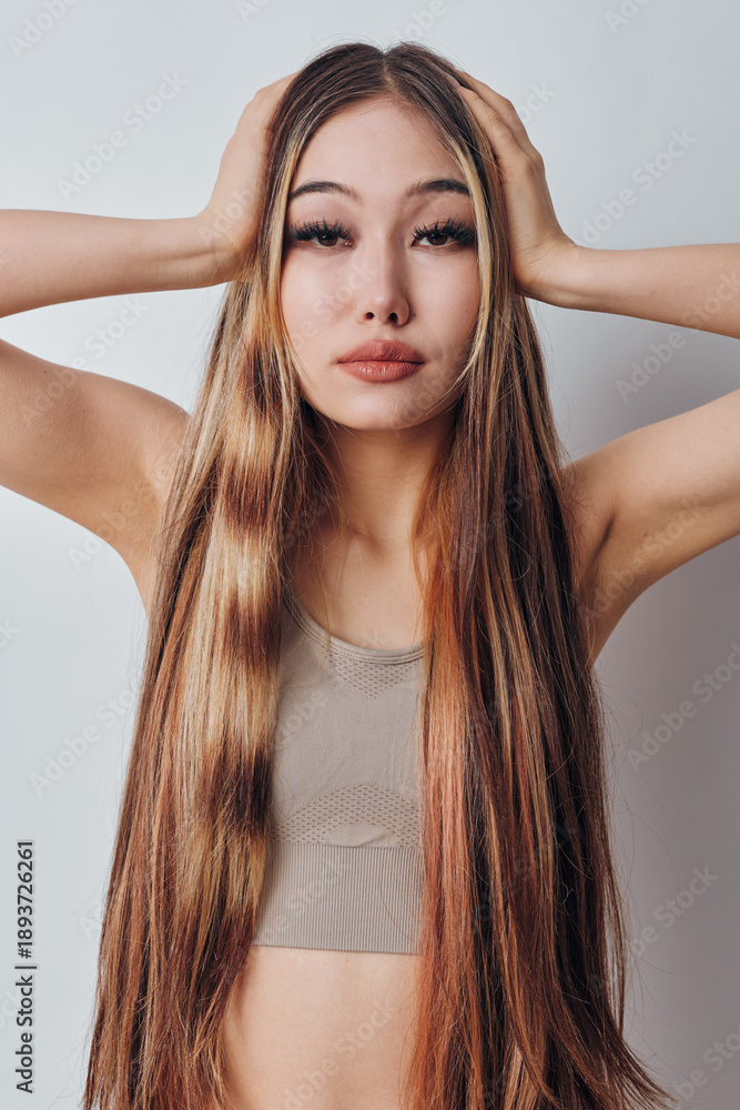 Fototapeta premium Woman with long straight hair posing with hands on head looking at camera wearing beige top in studio light on white background. Fashion, beauty and portrait closeup.