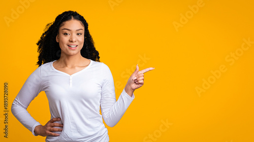 A young woman with curly hair smiles and points to the side. She wears a white long-sleeve shirt and stands against a bright yellow background. Her expression is friendly and inviting.