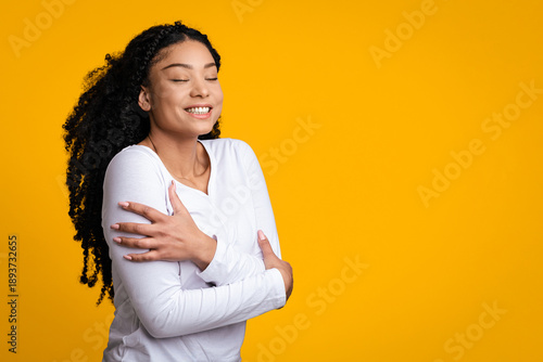 Φωτογραφία A Black woman with curly hair smiles while gently holding her arms around herself in front of a yellow background