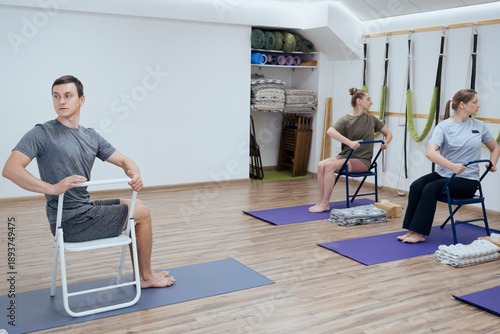 Tablou pe pânză Group of people doing seated spinal twist yoga asana with chair indoor