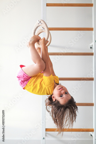Active little girl hanging upside down on gymnastic rings at home, smiling child in bright clothes enjoying indoor physical activity, childhood development, play healthy lifestyle concept