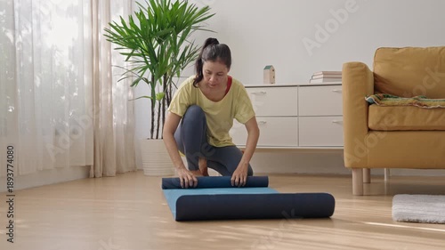 Asian Woman Rolling Up Yoga Mat After Practice, Discipline and Healthy Lifestyle Completion in Sunny Modern Apartment Morning Routine for Wellness