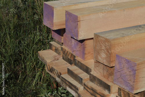 Stack of freshly cut wooden beams with visible end grain, neatly piled outdoors on grass, construction material prepared for building or carpentry work in a rural setting.
