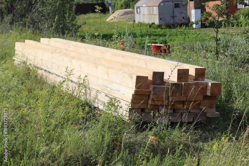 Large stack of wooden beams arranged in even rows outdoors, construction lumber stored on grass, natural building materials ready for use in residential or agricultural projects.