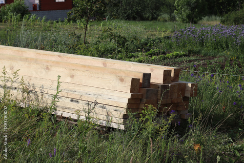 Large stack of wooden beams arranged in even rows outdoors, construction lumber stored on grass, natural building materials ready for use in residential or agricultural projects.