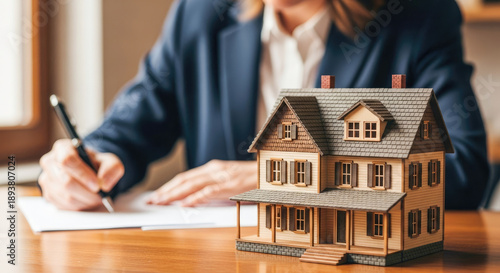 A woman in a suit writing on a piece of paper with a model house in front of her.