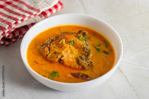 White bowl holds Indian katla/catla fish curry Bengali style with golden gravy, tender fish pieces, onion slices, and herbs, styled on marble surface with red cloth background.
