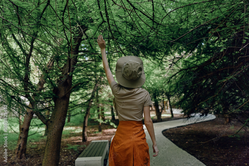 Obraz premium Woman in forest wearing a hat and orange skirt reaching up to tree branches on a winding path by a bench, exploring nature during a quiet summer walk in green woodland alone.