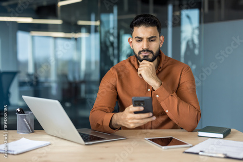 Young indian man in a contemporary office setting sitting at a wooden desk with a laptop, tablet, and documents, carefully reviewing information on his mobile phone, reflecting on business decisions