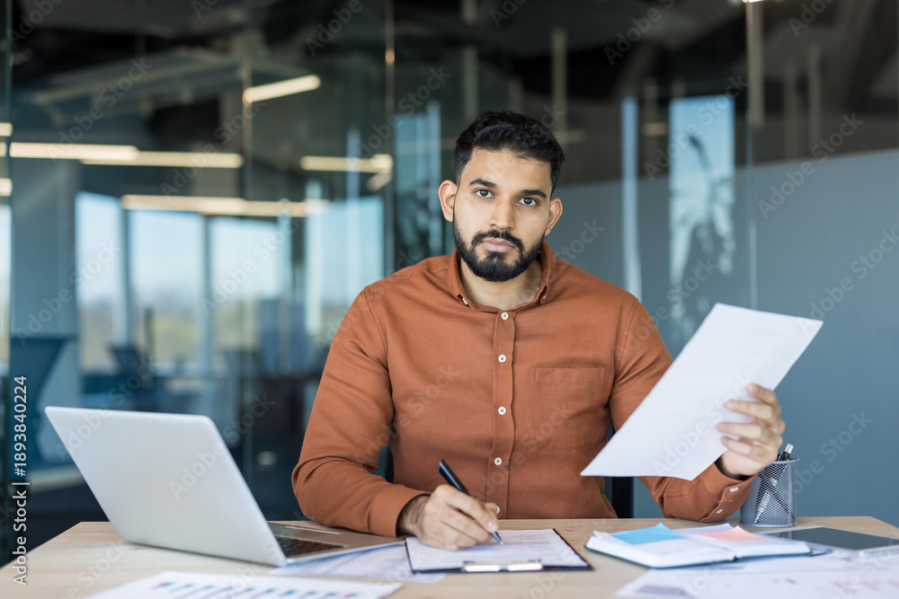 Obraz premium Young professional man sitting at an office desk, reviewing documents and writing on a clipboard, managing important business tasks with a laptop nearby