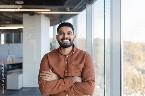 Young professional indian man smiling with crossed arms, standing confidently in a modern office with large bright windows, embodying success, leadership, and positive business attitude