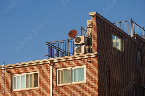 Close-up of an old antenna and outdoor air-conditioning units installed on the rooftop of a red brick residential building under a blue sky