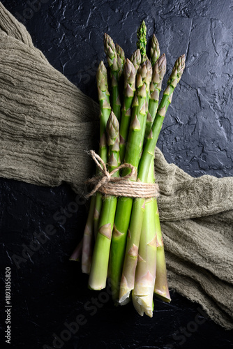 Bundled green asparagus tied with twine on kitchen cloth. Fresh raw vegetables shown on dark textured surface. Top view, flat lay