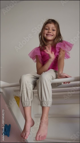 Cute Little Girl Smiles and Waves While Sitting on Climbing Frame: Playful Indoor Fun