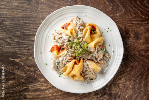 Photos A plate of creamy mushroom pappardelle pasta garnished with fresh herbs and a touch of red sauce, served on a white plate on a rustic wooden table