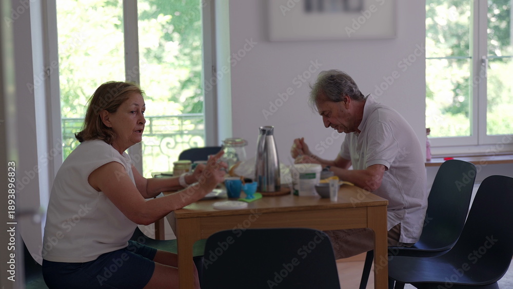 Obraz premium Senior couple eating breakfast together at home seated at dining table, enjoying morning meal with yogurt, coffee, and fruit near bright windows in cozy modern apartment
