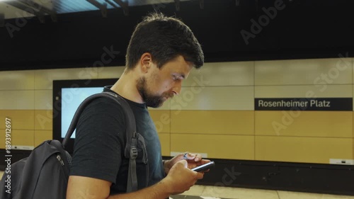 Man using smartphone while waiting on S-Bahn platform at Rosenheimer Platz station in Munich, Germany. Urban public transport scene, daily commute, modern transit infrastructure and city mobility