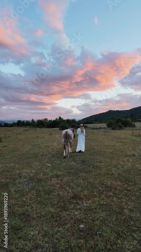 Girl walks with horse in field during sunset near mountains