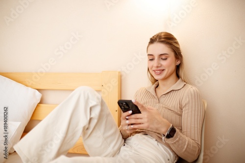 Beautiful smiling woman using her phone bedroom chair. A relaxing moment in a cozy bedroom.