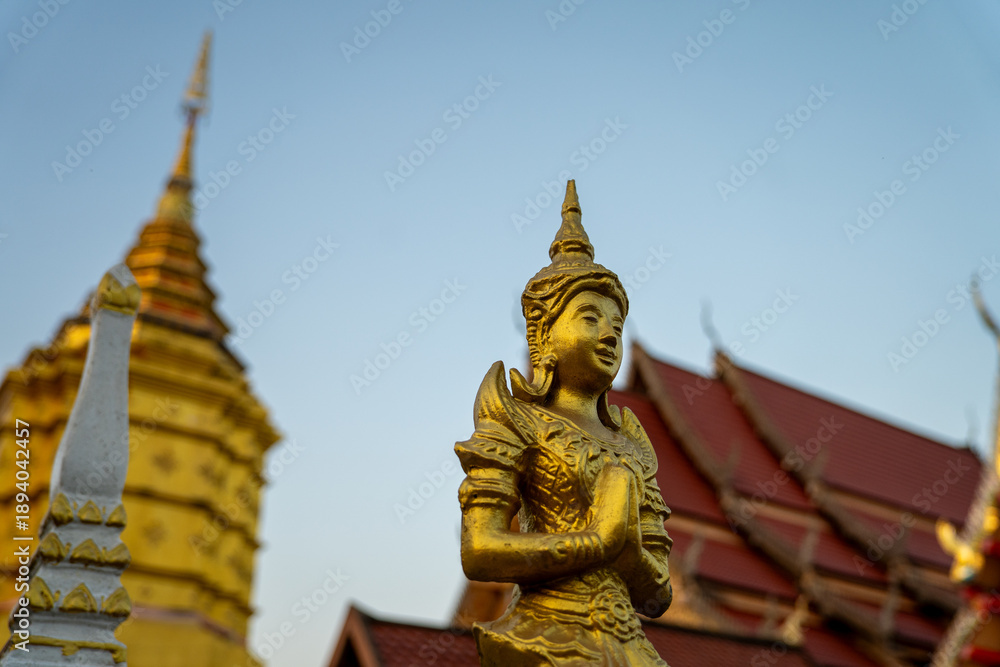 Fototapeta premium Golden angel statue in praying gesture with temple pagoda at sunset sky, sacred Thai sculpture representing faith, devotion, Wat Luang, Chiang Khong, Chiang Rai, Thailand