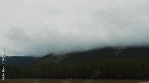 An atmospheric landscape of a mountain range wrapped in dense fog. Low clouds slowly drift across the slopes, hiding the peaks and creating a sense of mystery and depth. 